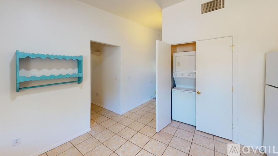A room with a white door, a white fridge, and a white wall with a blue shelf.