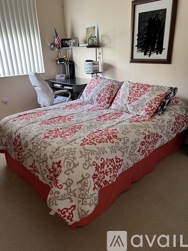 A bed with a red and white comforter is in the foreground of a room with a desk and chair.