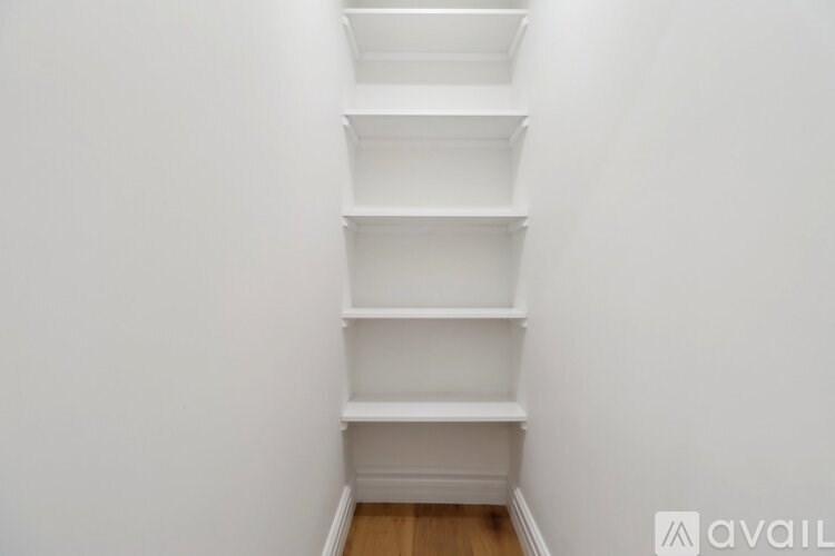 A white shelving unit with four shelves is in a corner of a room with white walls and wooden flooring.
