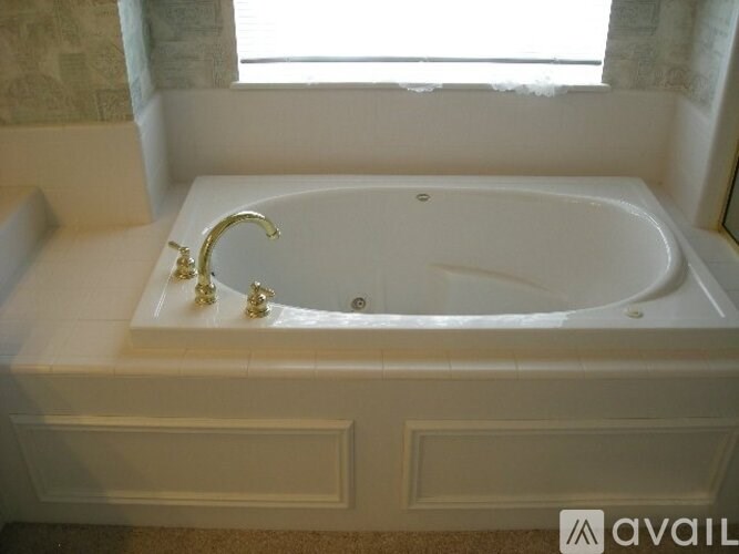A white bathtub with gold faucets is in a bathroom with a window.