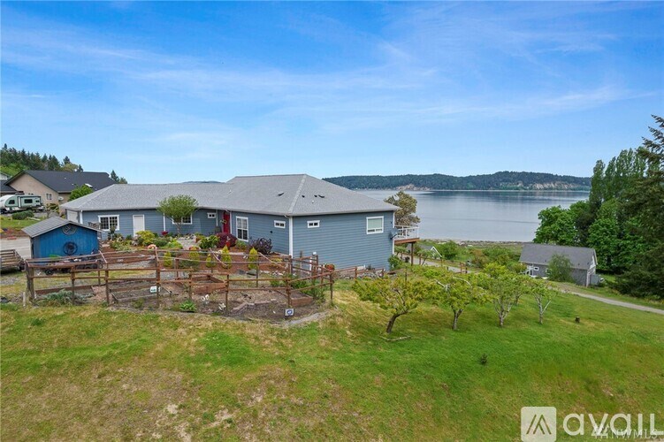 A house with a blue roof is situated near a body of water with a fenced yard in front.