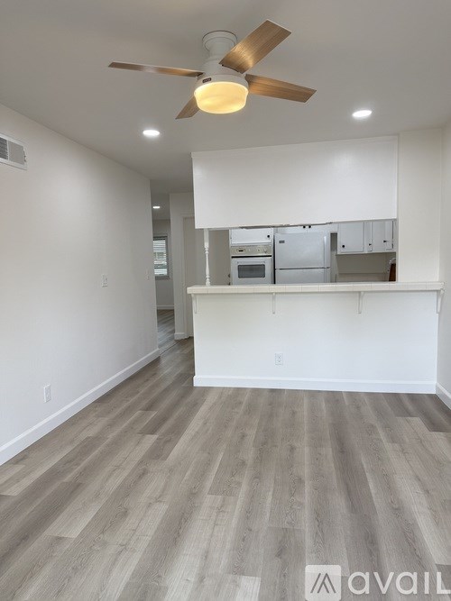 A room with a ceiling fan and a kitchen area in the background.