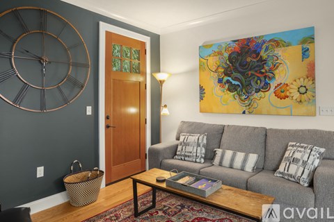 A living room with a grey couch, a large clock on the wall, and a painting on the wall.