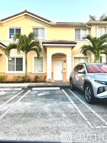 A white car is parked in a parking lot in front of a yellow house.