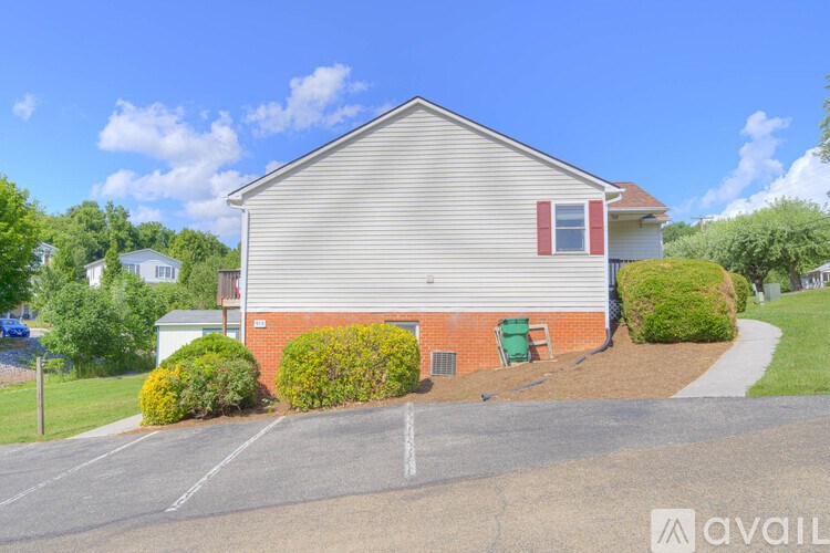 A house with a grey roof and red shutters is surrounded by greenery and a parking lot.