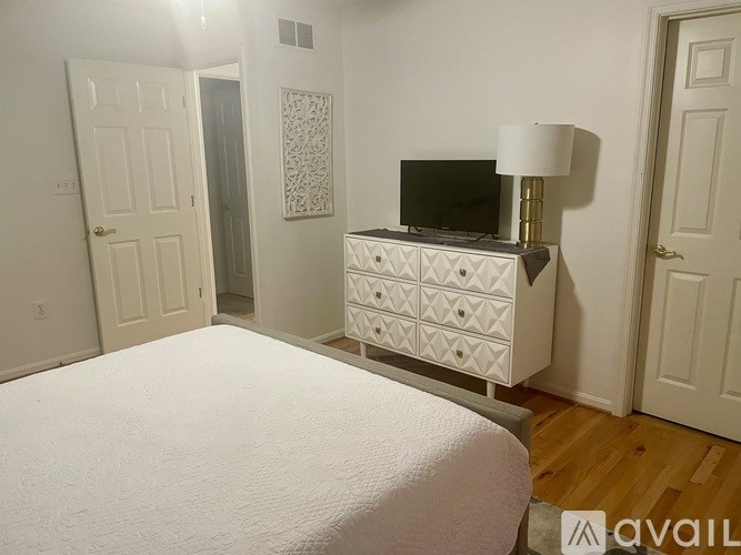 A bedroom with a white bed, a white dresser with a black television on top, and a white lamp.