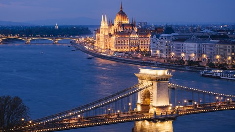The image shows a beautiful view of Budapest with the Hungarian Parliament building and the Chain Bridge illuminated at dusk.
