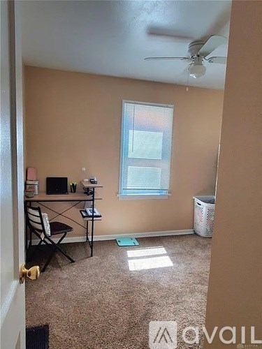A room with a fan, a desk with a laptop and a chair, a window with blinds, and a trash can.