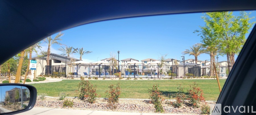 A car's side mirror shows a view of a building complex with a fence and greenery.