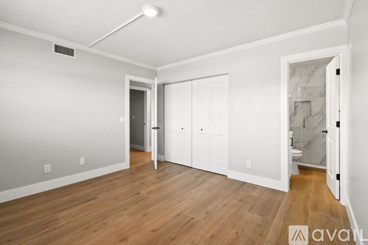 A room with wooden floors and white walls, with a door on the left and a bathroom visible through the door.