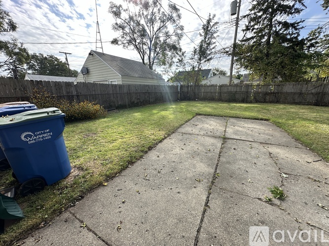 A backyard with a blue trash bin and a concrete path.