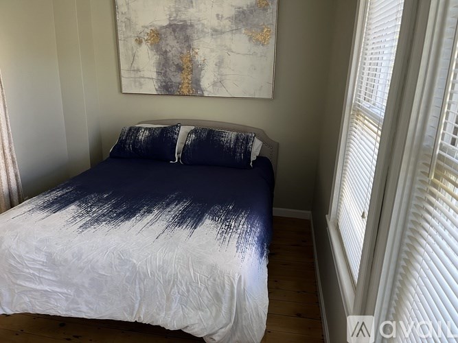 A bed with a blue and white comforter is in a room with a painting on the wall and a window with blinds.