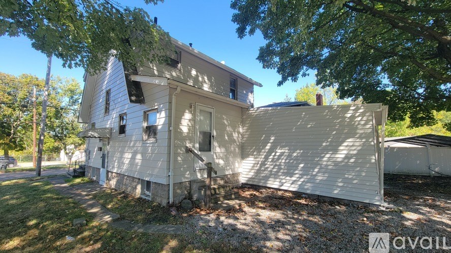 A house with a white exterior is surrounded by trees and has a gravel area in front.