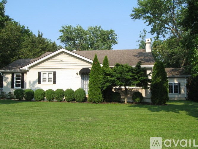 A white house with a brown roof and a chimney is surrounded by green trees and bushes.