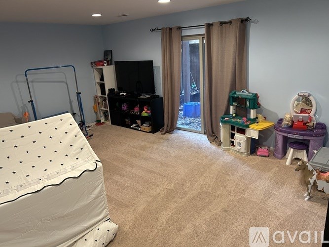A room with a bed covered in a white sheet with black dots, a TV, a chair, a table, a window with curtains, and a few toys scattered around.