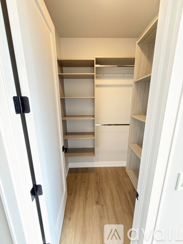 A white door with a black handle is open to reveal a walk-in closet with shelves and a wooden floor.