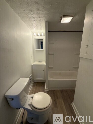 A white toilet in a small bathroom with wood flooring and a white bathtub.