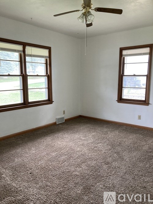 A room with a carpeted floor, a ceiling fan, and two windows.