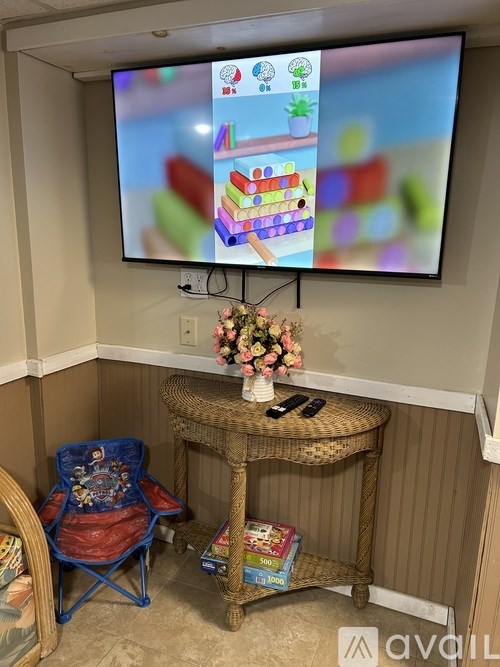 A television screen shows a colorful children's game on a wall above a small table with a blue chair and a basket of flowers.