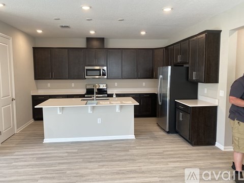 A man is standing in a kitchen with a refrigerator, microwave, and oven.