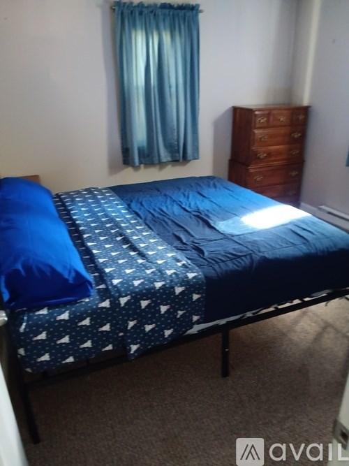 A bed with a blue comforter and a blue pillow is in a room with a window covered by a blue curtain.