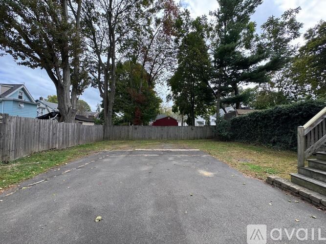 A paved driveway leads to a red garage with a wooden fence and trees in the background.