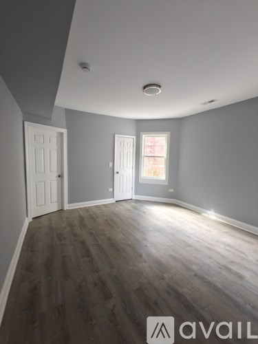 A room with grey walls and wooden flooring with a door on the left and a window on the right.