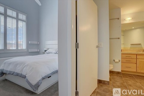 A bedroom with a bed, a window with blinds, and a door leading to another room.