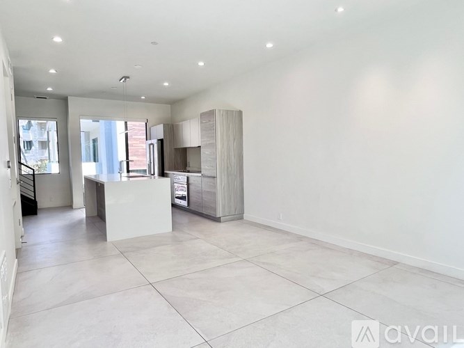 A spacious, empty room with white walls and flooring, and a kitchen area with cabinets and a countertop.