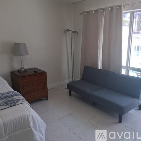 A grey couch sits in a living room with a lamp and a bed in the background.