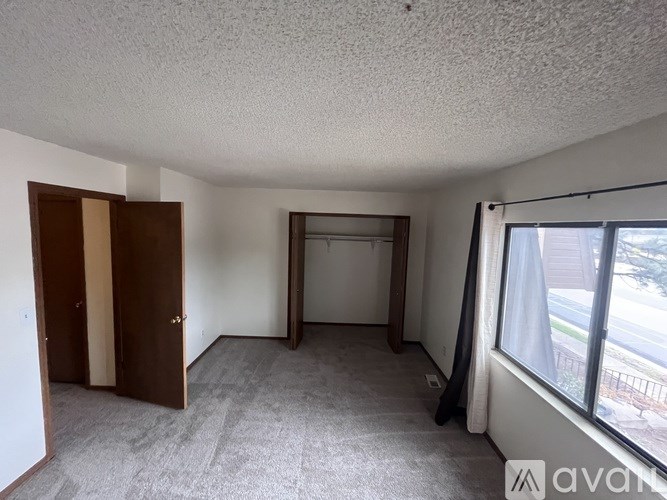 A room with a carpeted floor, a window with a view of the outside, and a door on the left side.