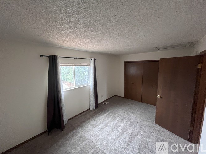 A room with a carpeted floor, a window with curtains, and a wooden wardrobe.