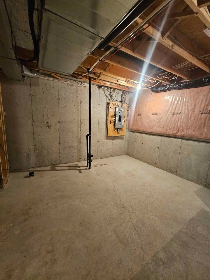 A room with a concrete floor and walls, a wooden beam on the ceiling, and a black pipe running along the wall.