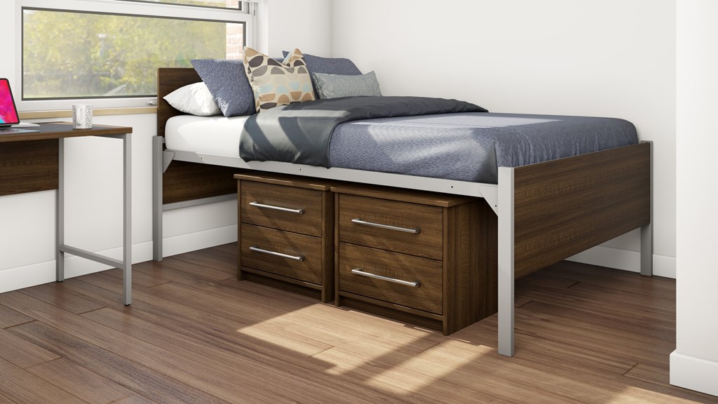 A bed with a grey sheet and pillows is in a room with a desk and drawers.