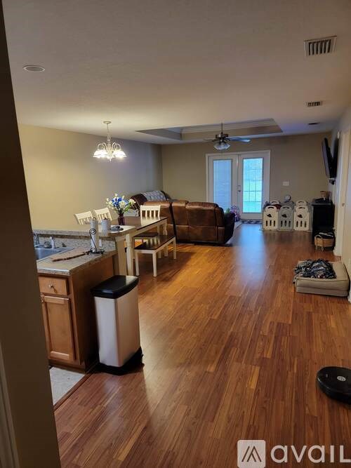 A living room with a brown couch and a kitchen area with a sink and a trash can.