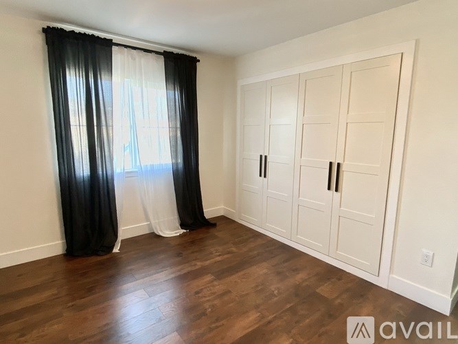 A room with a large window covered by black and white curtains and a set of white cupboards.