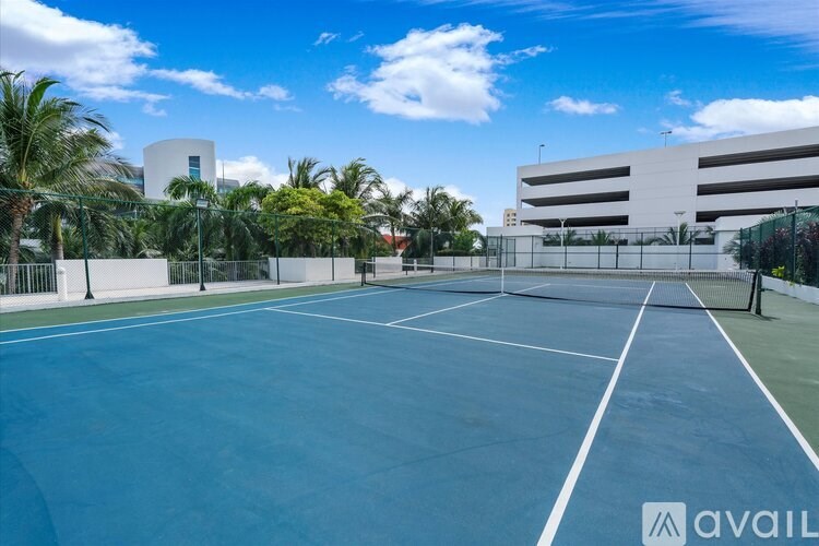 A tennis court with a blue surface and white lines, surrounded by a fence and palm trees.