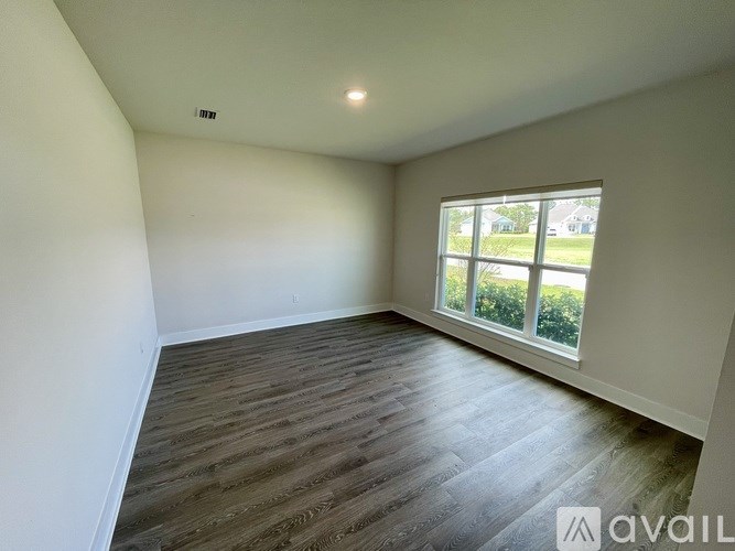 A room with wooden flooring and a window showing a view of a green lawn and a house.