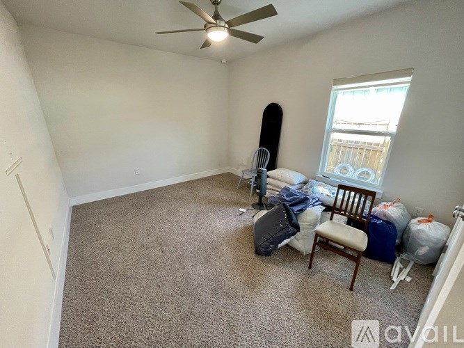 A room with a carpeted floor, a ceiling fan, a chair, and a window with a view of a fence and trees outside.