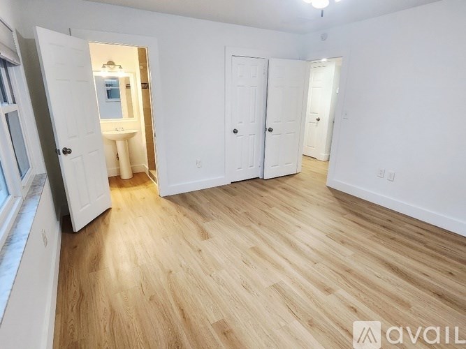 A room with wooden flooring and white walls, with a door open to a bathroom.