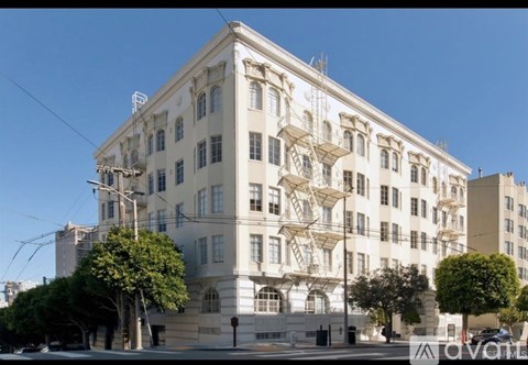 A large white building with many windows and balconies is surrounded by trees and has a clear blue sky above it.