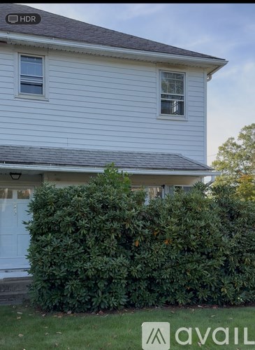A house with a white siding and a grey roof with a hedge in front.