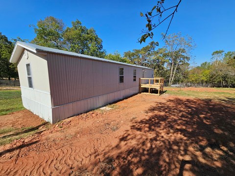 A small white building with a brown roof is situated on a dirt area.