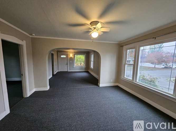 A spacious room with a carpeted floor, a ceiling fan, and a view of the outside through the windows.