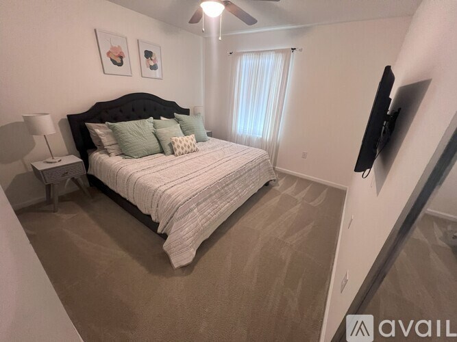 A bedroom with a bed, a nightstand, a lamp, a window with curtains, and a flat-screen TV mounted on the wall.