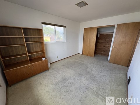 A room with a carpeted floor, a wooden bookshelf, and a window with blinds.