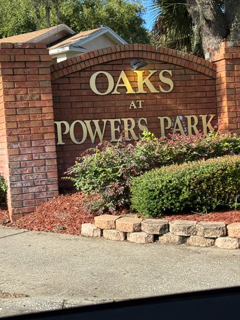 A sign that says "Oaks at Powers Park" is in front of some bushes.