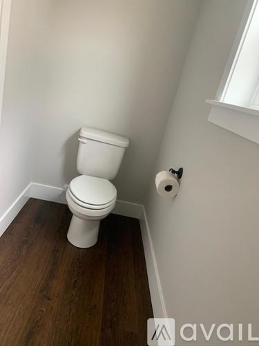 A white toilet sits in a small bathroom with wood flooring and a window.