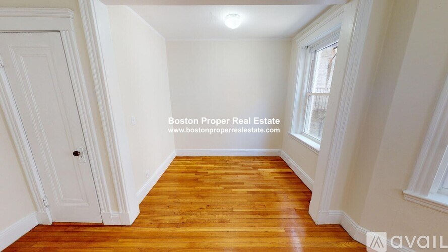 A room with wooden flooring and white walls, with a door on the left and a window on the right.