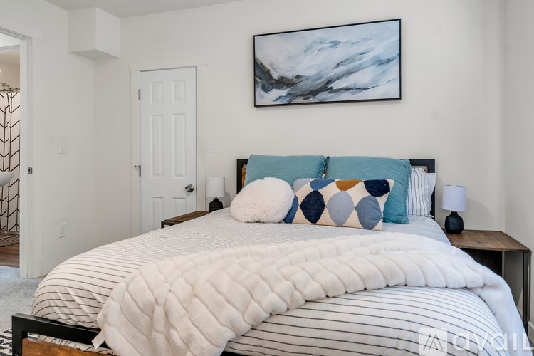 A bed with a white and blue comforter and pillows is in the foreground of a room with a painting on the wall.